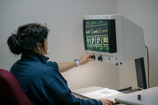 Person sitting at a desk using a CCTV magnifier device to view printed text on a monitor, adjusting the control knob for better visibility